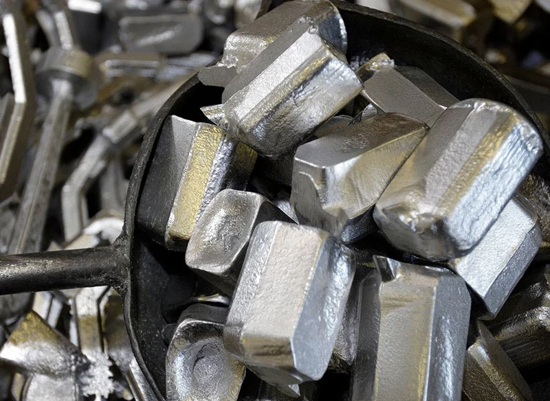 Close-up of shiny, rectangular silver metal ingots piled in a container, with a reflective surface. The scene conveys industrial precision and abundance.